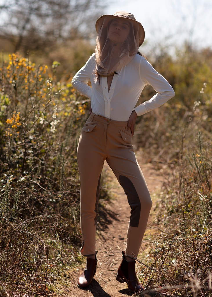 Female model in beige riding breeches, white shirt and a safari hat standing in a field of grass