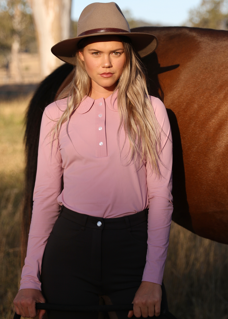 Young woman in long hair, hat and a pink blouse leaning against a brown horse