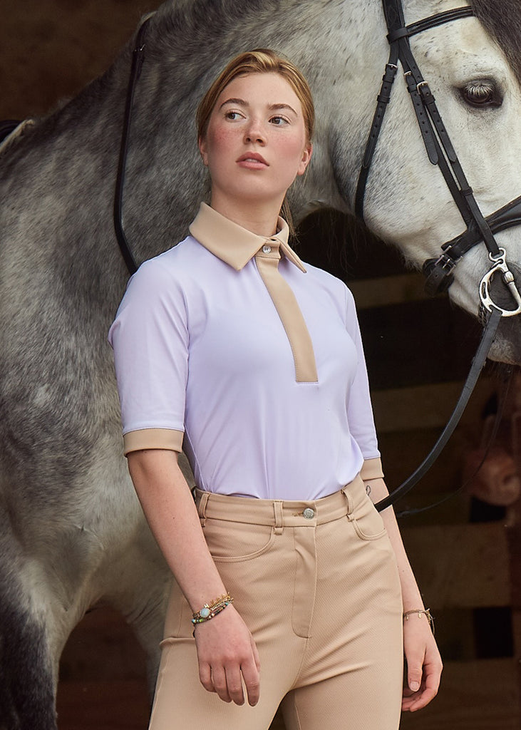 Young woman in lilac top and beige breeches standing in front of grey horse 
