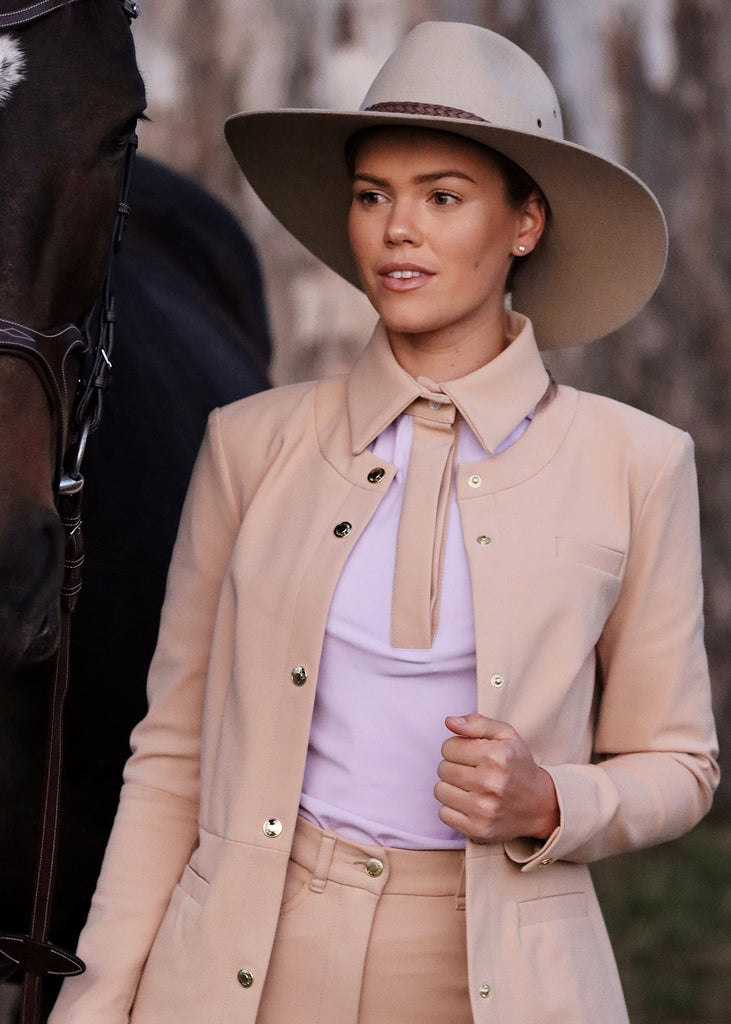 Young woman dressed in a beige hat, jacket and breeches next to a brown horse