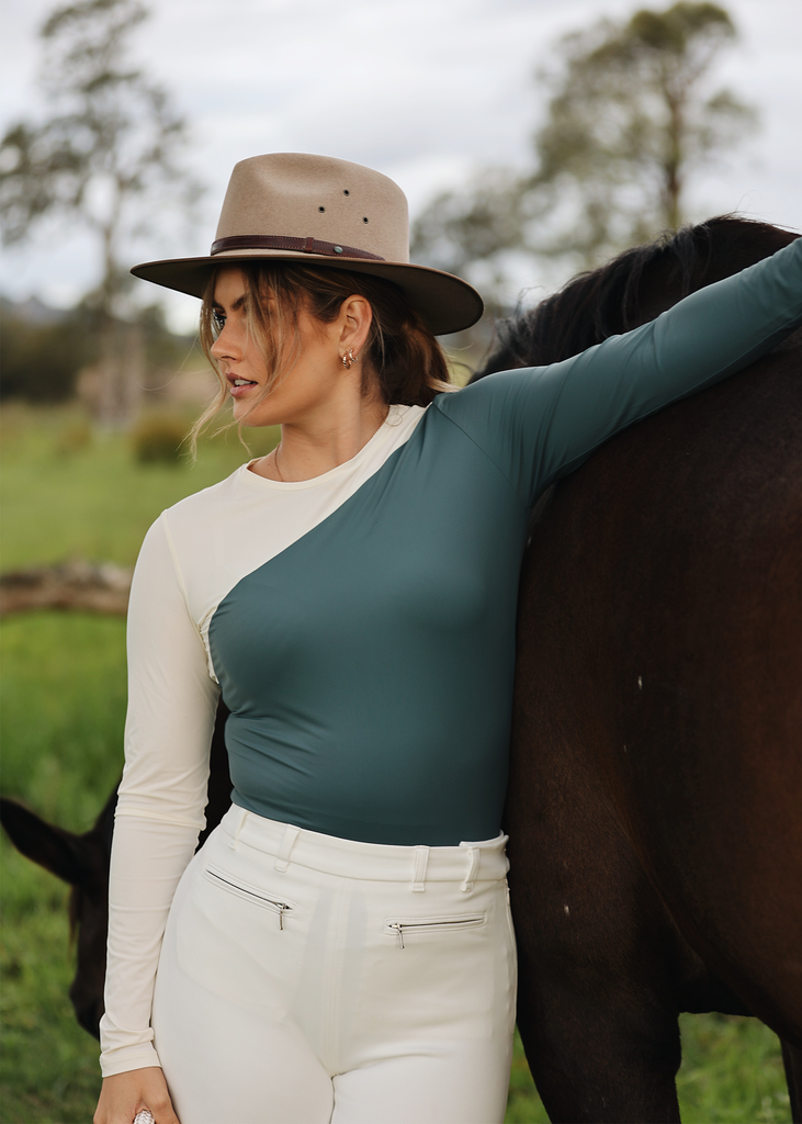 Young woman in a hat leaning against Wearing a brown horse. and color blocked top with white riding breeches