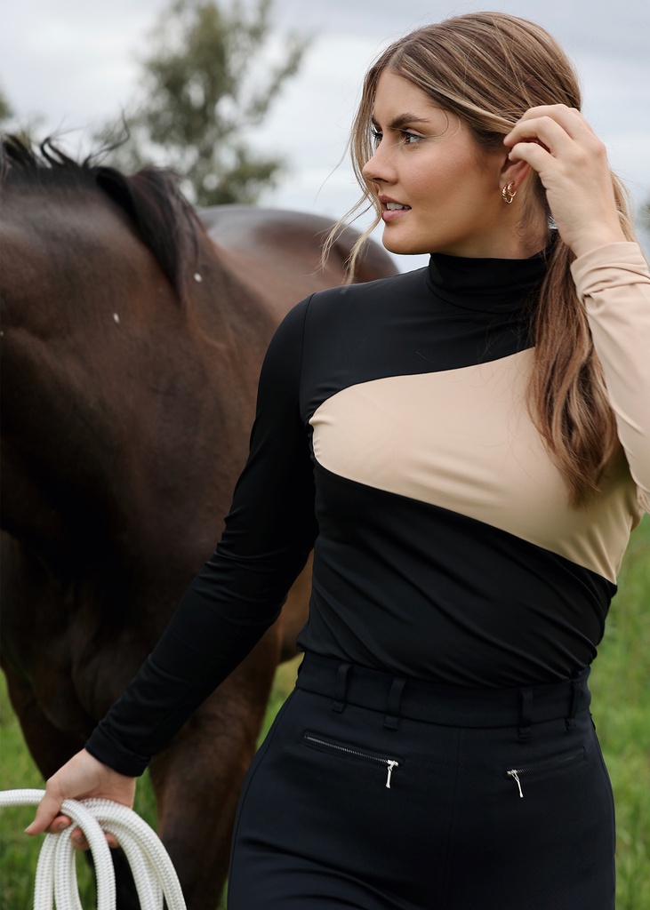 Woman with long brown hair walking with a horse. Wearing a black and tan colorblock turtle neck top. 