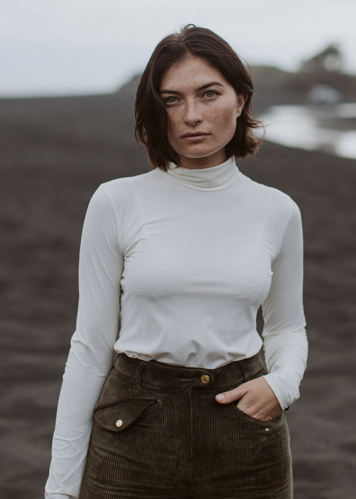 Dark haired woman standing on a empty field looking in to the camera. Wearing a white Lycra roll neck top and corduroy breeches. 