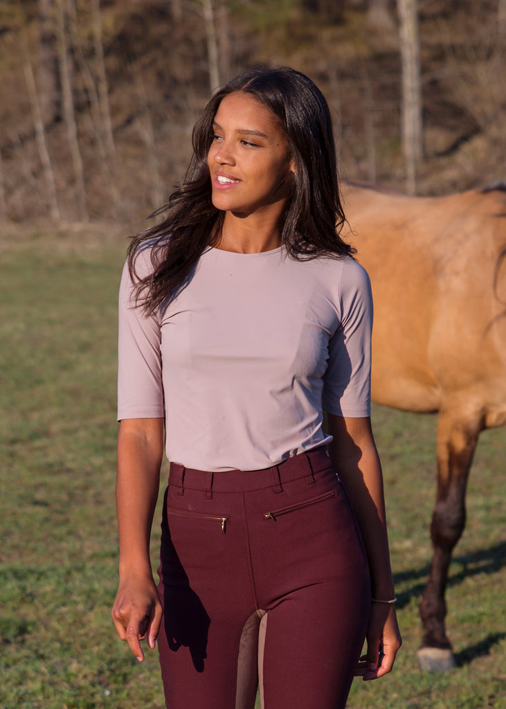 Smiling woman with a palomino horse in the background. She is wearing a lilac t-shirt with dark red breeches.