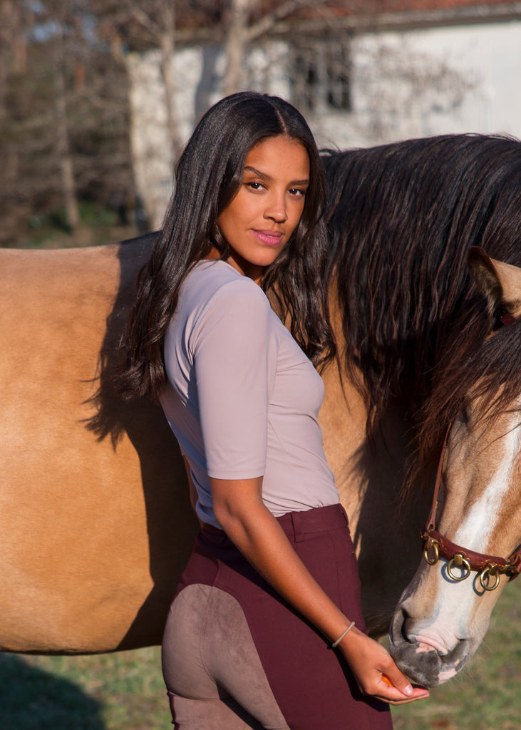 Woman petting her horse, glancing over her shoulder in to the camera. Wearing a lilac slim fit t-shirt and dark red full seat breeches. 