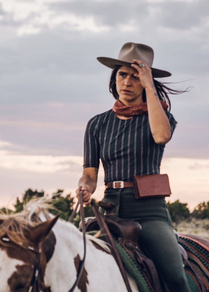 Cowgirl style woman in a hat riding her horse. Wearing a scarf around her neck, a striped t-shirt, brown leather belt bag and dark green breeches. 