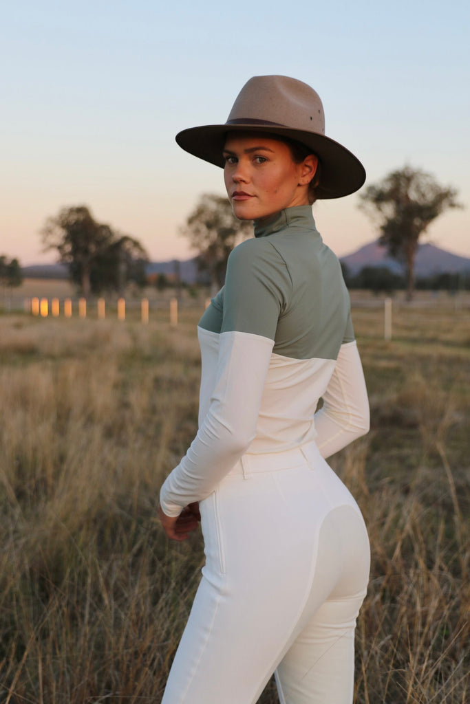 Woman in hat standing on a field looking over her shoulder wearing a white/green turtle neck and white breeches 